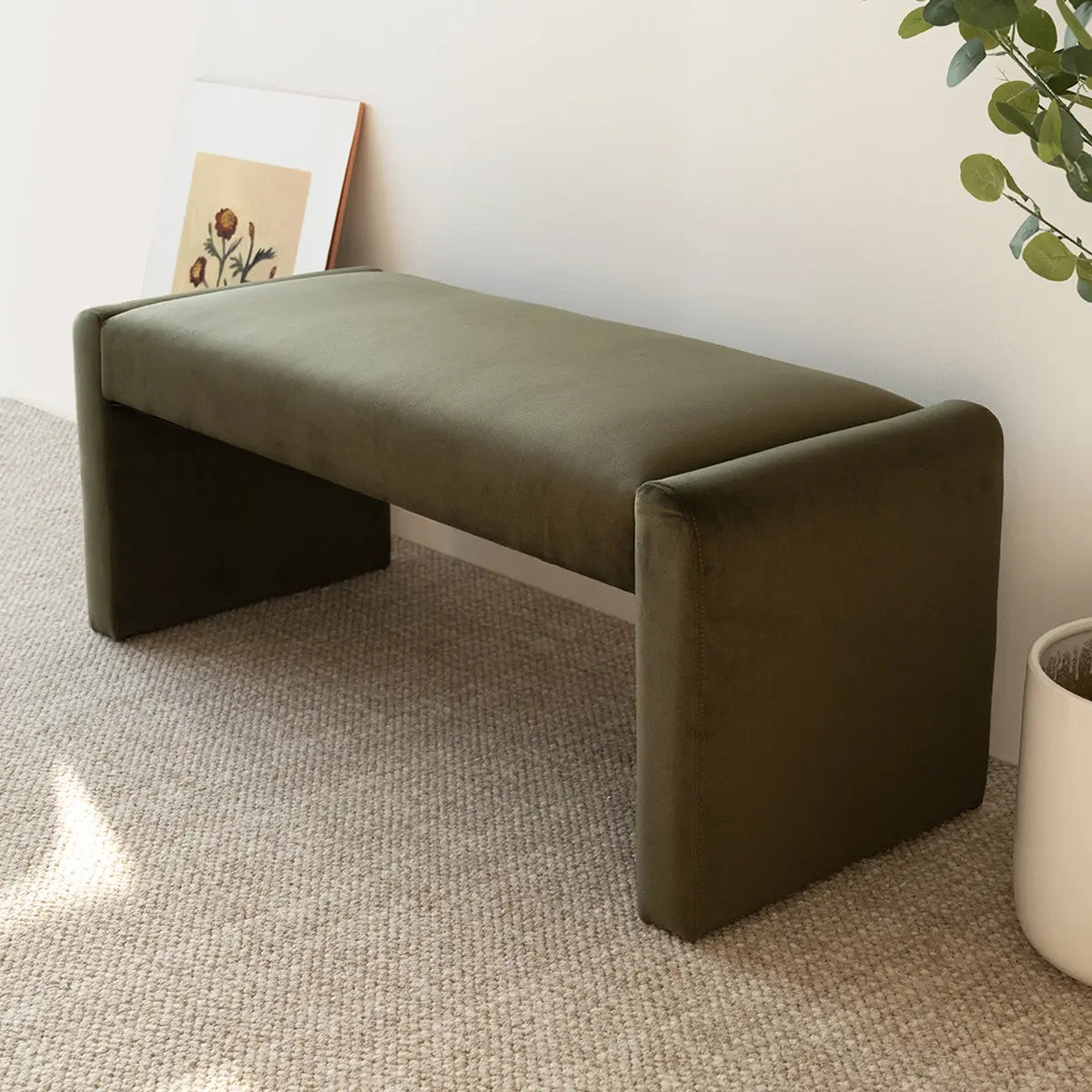 Kaia bench with green velvet, beige carpet, white wall, and plant in modern living room setting.