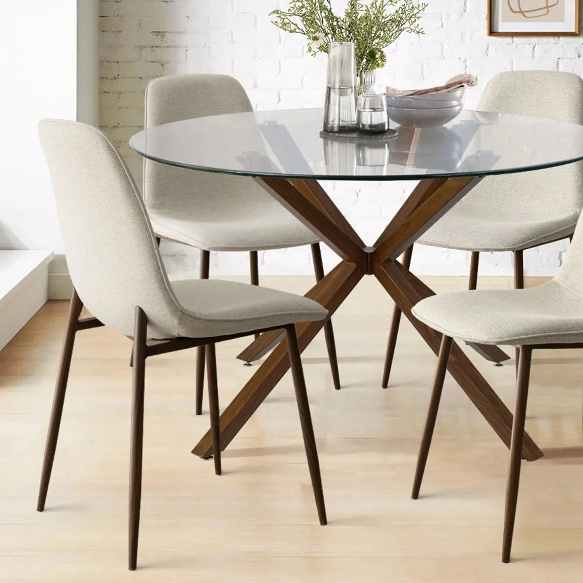 Oliver & Oslo Glass Round Dining Table Set with beige chairs, wooden legs, white brick wall, wood floor.