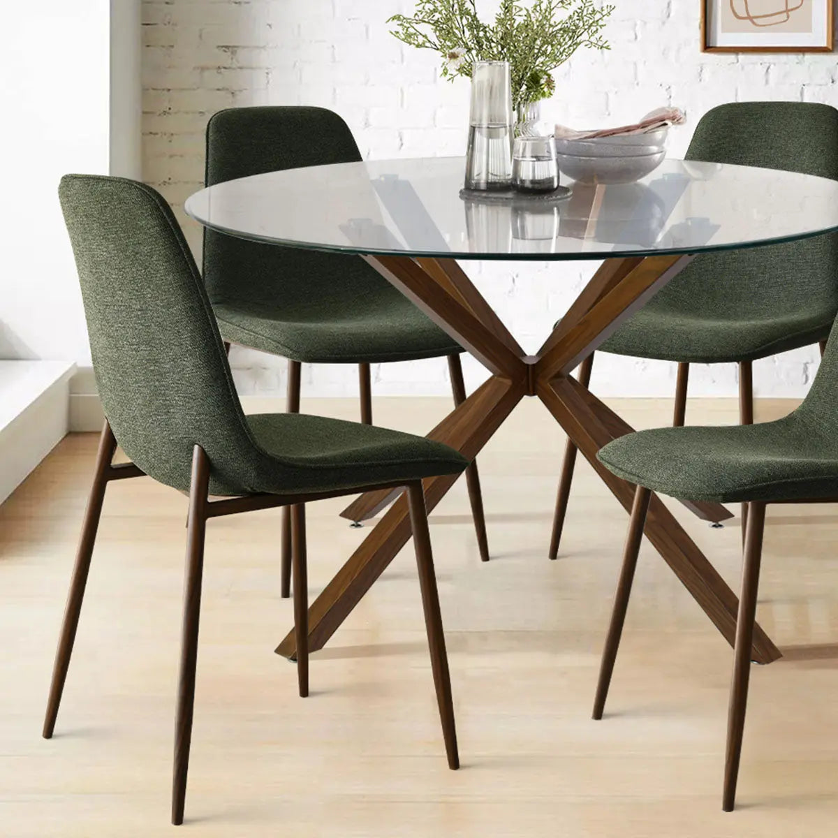 Oliver & Oslo dining set, glass round table, green chairs, wood legs, brick wall background, light flooring.